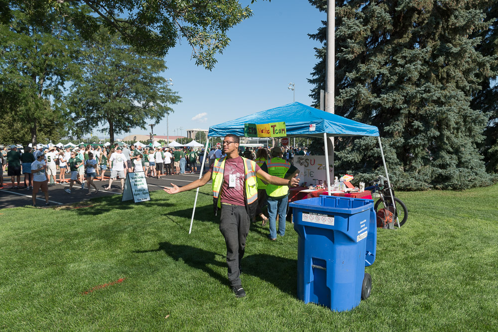 A student with a safety vest walks towards a recycling bin on a grassy field at a CSU athletic event.